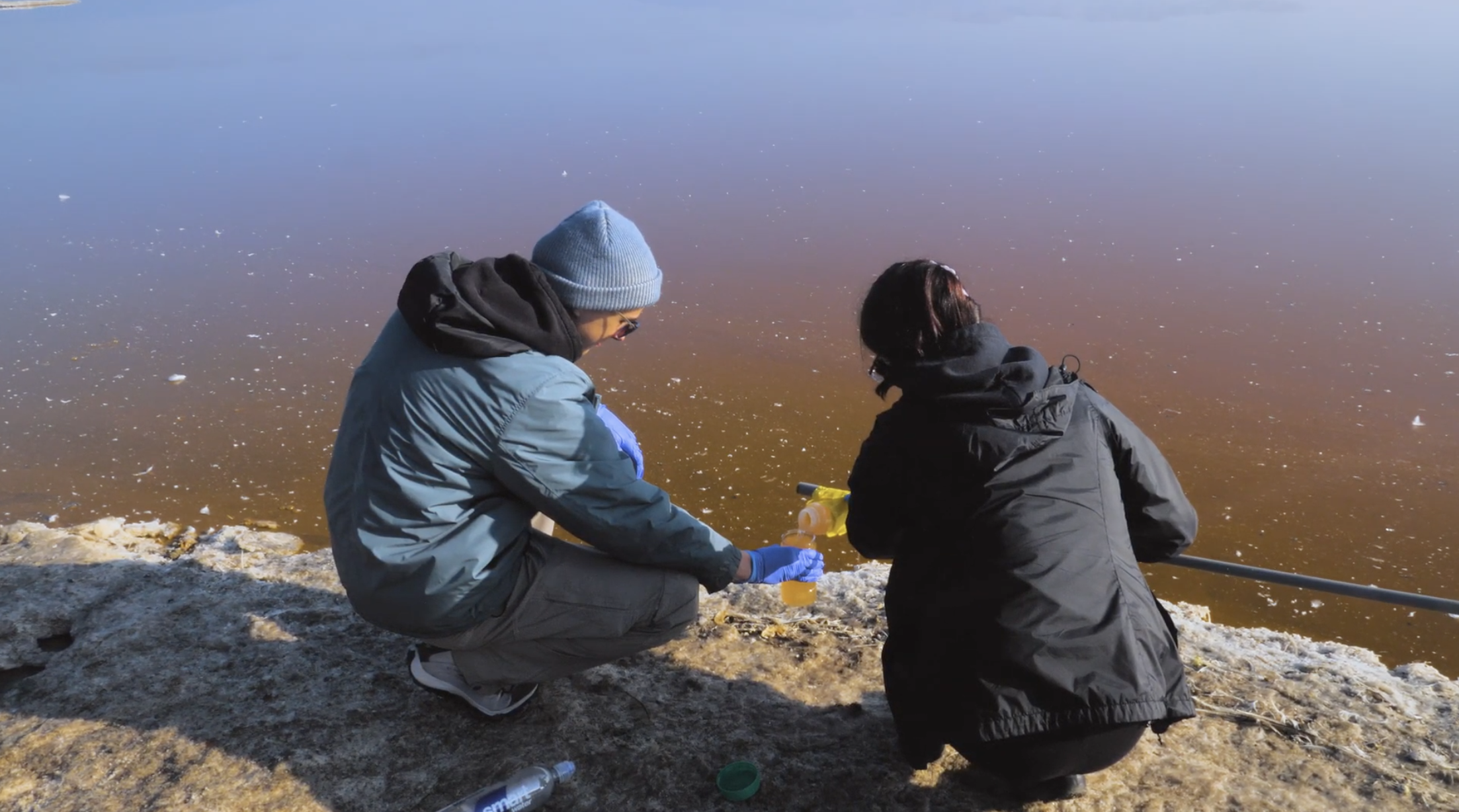 Two students kneel to sample brown-colored water, standing on a salt flat.
